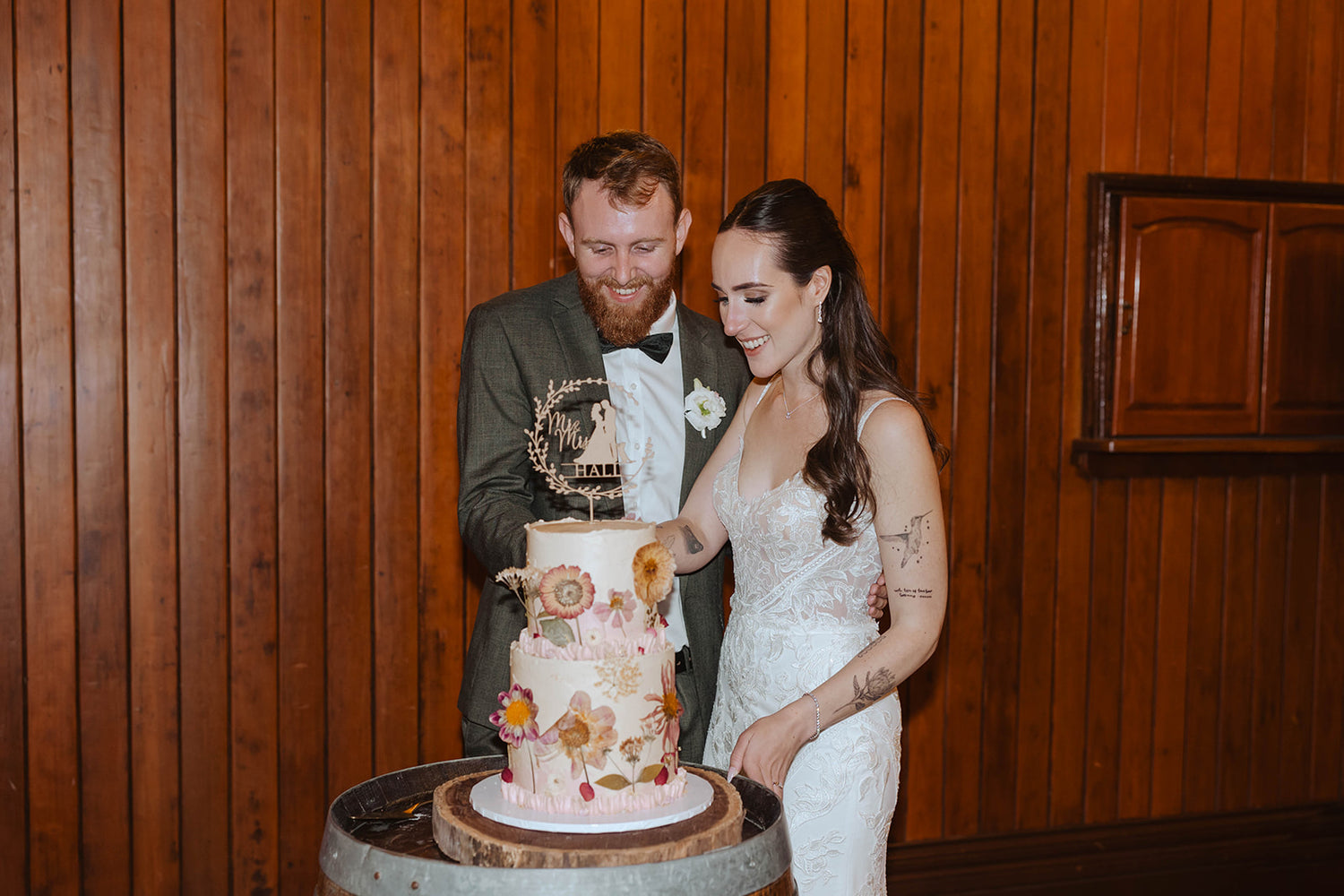 Happy bride and groom cutting a two-tier rustic wedding cake with buttercream icing, pressed flowers, and a personalized topper, captured at a Brisbane celebration by Caldera.CAKING.