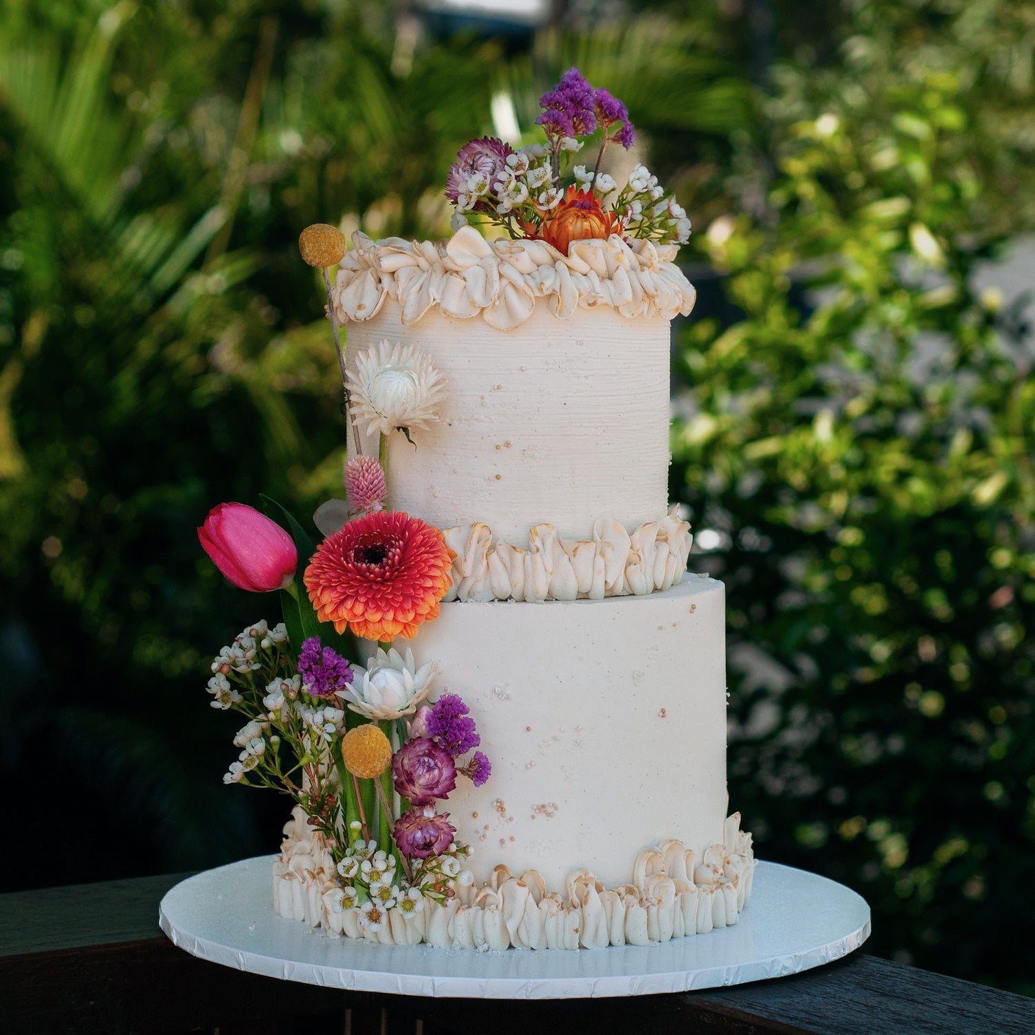 Rustic two-tier buttercream wedding cake adorned with fresh wildflowers, including tulips, daisies, and bright orange blooms, crafted for Brisbane weddings by Caldera.CAKING.