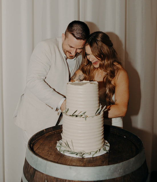Smiling bride and groom cutting a two-tier wedding cake with a rustic buttercream finish and fresh greenery accents, captured at a Brisbane celebration by Caldera.CAKING.