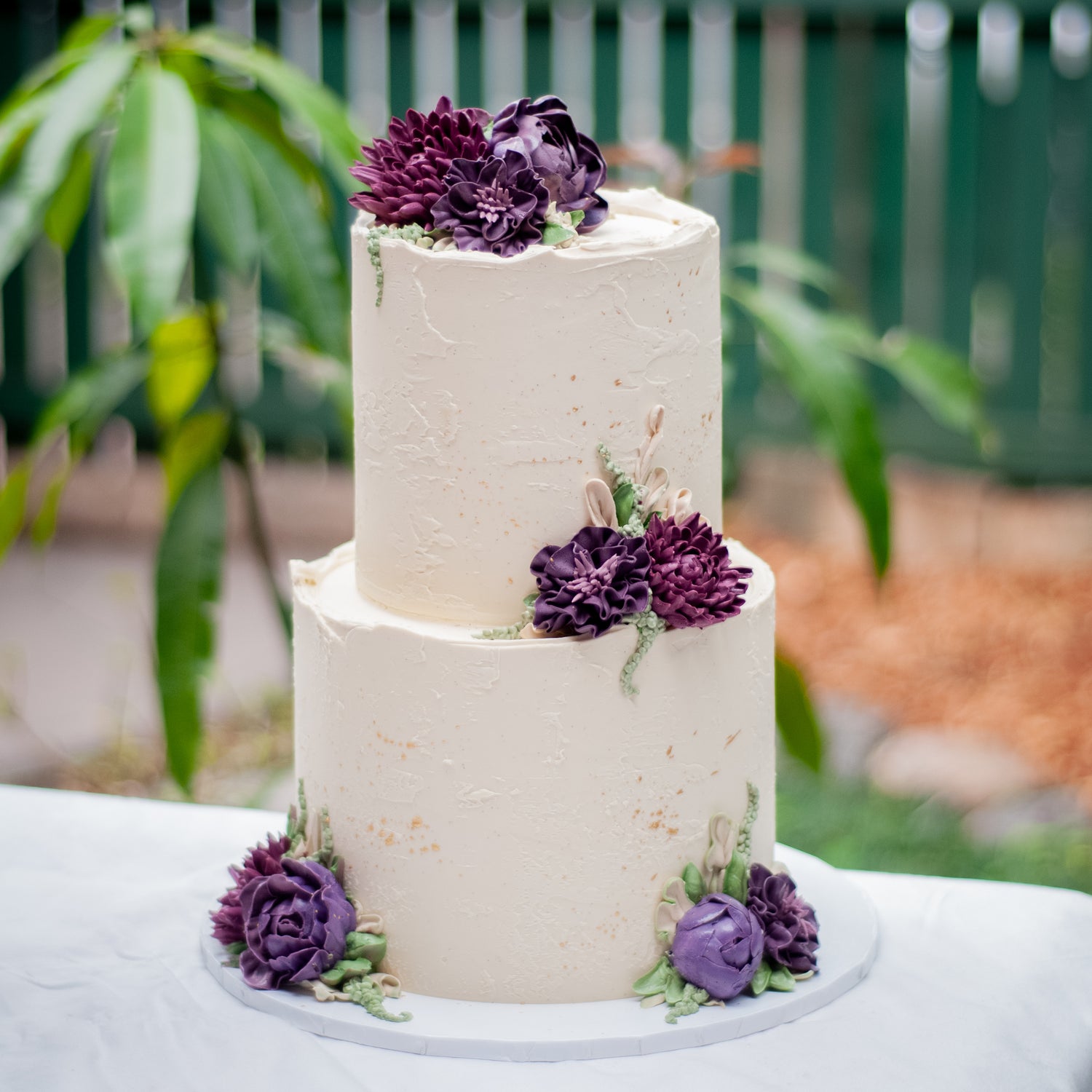 Two-tier buttercream wedding cake adorned with deep purple flowers and smooth icing, ideal for sophisticated Brisbane wedding celebrations.